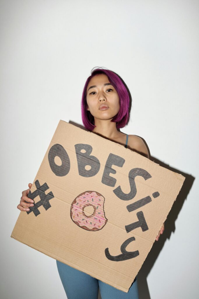 Asian woman with purple hair holding a hand-drawn obesity sign in a studio setting, promoting awareness.