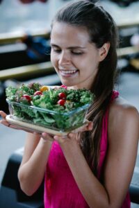 Smiling woman in a pink top holding a bowl of fresh salad with cherry tomatoes and greens indoors.
